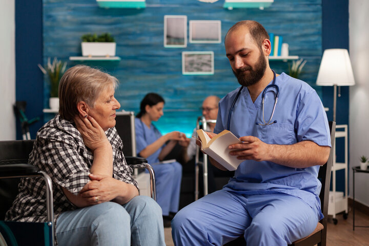 care worker in a nursing home reading a book for a woman in a wheelchair