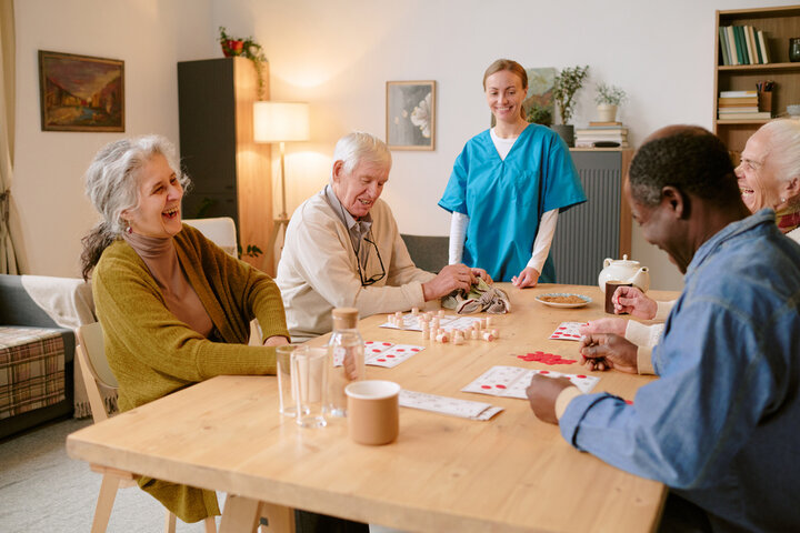 Wide shot of young nurse checking residents of nursing home sitting at wooden table and playing bingo game